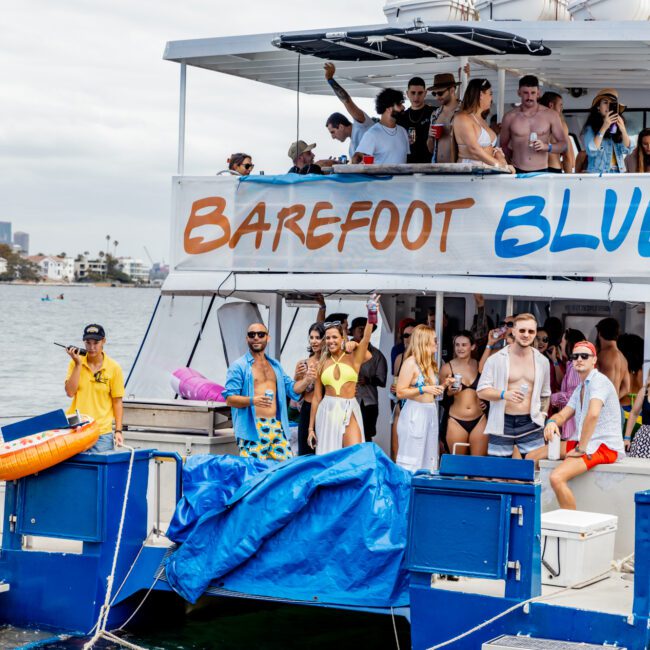 A lively group of people enjoys a yacht party on the "Barefoot Blue" boat. Dressed in summer outfits, some holding drinks, they appear to be dancing and socializing. The yacht, part of The Yacht Social Club Sydney Boat Hire, is docked on a body of water, creating a festive atmosphere.