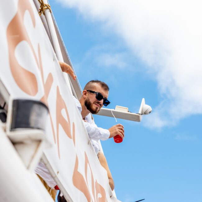 A man with a beard and sunglasses, wearing a white shirt, leans over the side of a boat holding a red cup. The boat, part of The Yacht Social Club Sydney Boat Hire, has a large banner with partial text visible. The sky is bright blue with a few clouds.