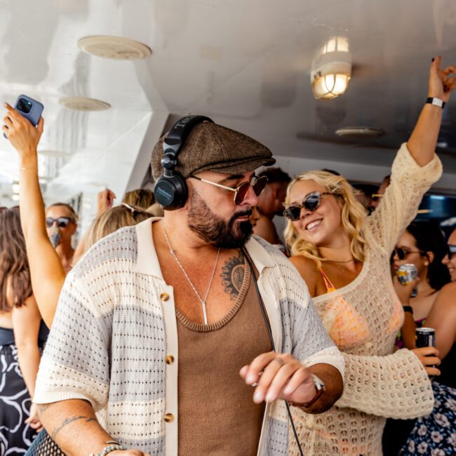 A man wearing a cap and sunglasses is DJing at an outdoor party on a boat from The Yacht Social Club Sydney Boat Hire. He has headphones on and is using a mixing console. People around him are dancing and enjoying the music, including a woman with blonde hair in a beige outfit, raising her arm joyfully.