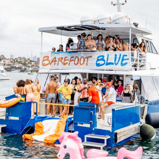 A double-deck boat named "Barefoot Blue" is packed with people enjoying a Sydney Harbour Boat Hire party by The Yacht Social Club. The deck is filled with partygoers, and some are swimming near the boat. In the water, there are large inflatable pink unicorn pool floats. The sky is overcast.