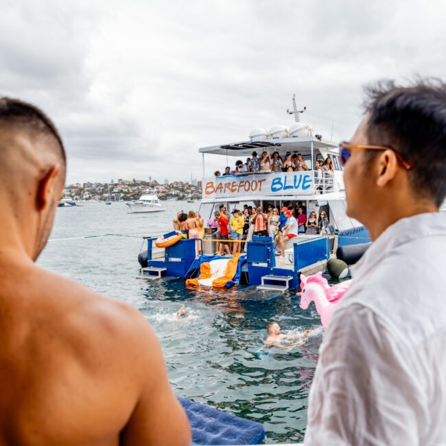 A group of people are enjoying a boat party on a cloudy day. The Yacht Social Club Event is lively with several individuals swimming near a boat named "Barefoot Blue." In the foreground, two men, one shirtless and the other in a white shirt, observe the festivities.