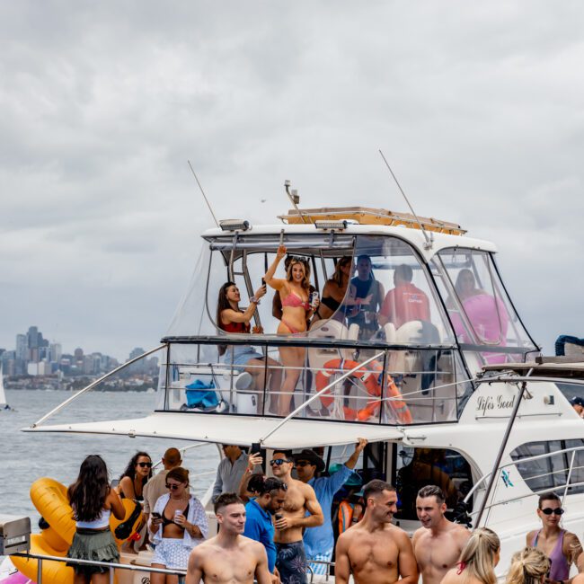 A lively scene on a two-tier yacht with young adults enjoying a party. People are dancing, talking, and holding drinks. Some are in swimwear under a cloudy sky. In the background, a city skyline and other boats are visible. Experience this with The Yacht Social Club Sydney Boat Hire for luxurious boat parties in Sydney!