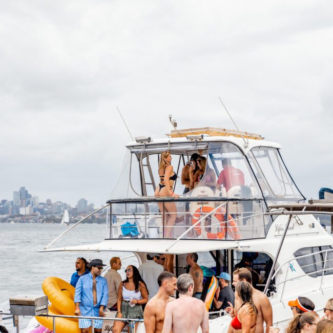 A group of people are gathered on a yacht on a cloudy day. Some individuals are at the front of the boat, while others are on the upper deck. The boat is decorated with various pool floats and party items. The city skyline and water are visible in the background, courtesy of Luxury Yacht Rentals Sydney.