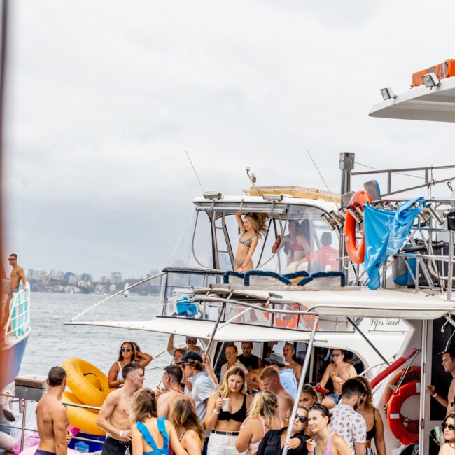 A crowded boat party is captured with people socializing, drinking, and enjoying the event. The boat is anchored in Sydney Harbour, and the sky above is overcast. Some people are on the top deck, while others gather on the main deck. The scene exudes a lively and festive atmosphere, typical of Boat Parties Sydney The Yacht Social Club.