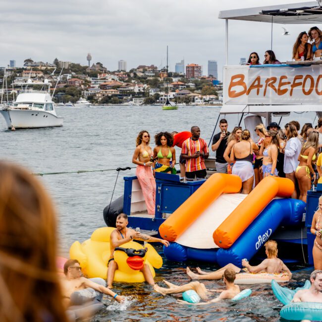 A lively boat party on a cloudy day features people enjoying themselves on an anchored boat named "BAREFOOT." Several attendees are on the deck, while others are in the water, some using a waterslide and an inflatable duck float. Other boats and a city skyline add to the charm of Boat Parties Sydney The Yacht Social Club.