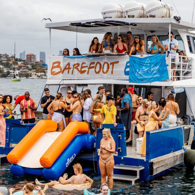 A lively group of people enjoys a boat party hosted by The Yacht Social Club. Some are on the deck with the name "BAREFOOT" visible, while others swim or float on pool inflatables in the water, with a slide extending from the boat. Everyone appears to be having a fun and festive time.