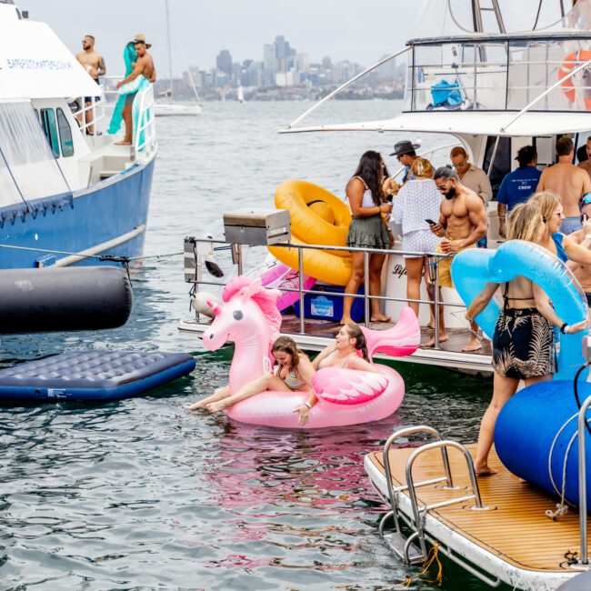 An outdoor scene with people enjoying a boat party. Two large boats are docked side by side, provided by The Yacht Social Club Sydney Boat Hire. Some people are on the boats, while others are in the water on inflatable floats, including a pink unicorn. A city skyline under a cloudy sky forms the background.