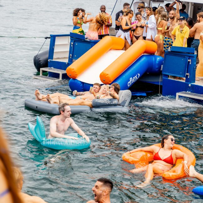 People having fun on inflatable floats in the water near a boat named "Barefoot," courtesy of Boat Rental and Parties Sydney The Yacht Social Club. Some are sliding into the water from a slide attached to the boat. A crowd is gathered on the boat's deck, with a cityscape visible in the background.