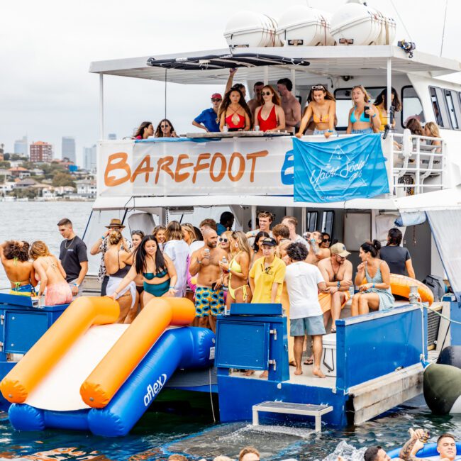 A lively scene of a boat party shows people enjoying themselves on a two-deck vessel named "Barefoot" from Luxury Yacht Rentals Sydney. Guests in swimsuits are seen on the boat, while others float on inflatables in the water. The boat features slides, with a city skyline in the distant background.