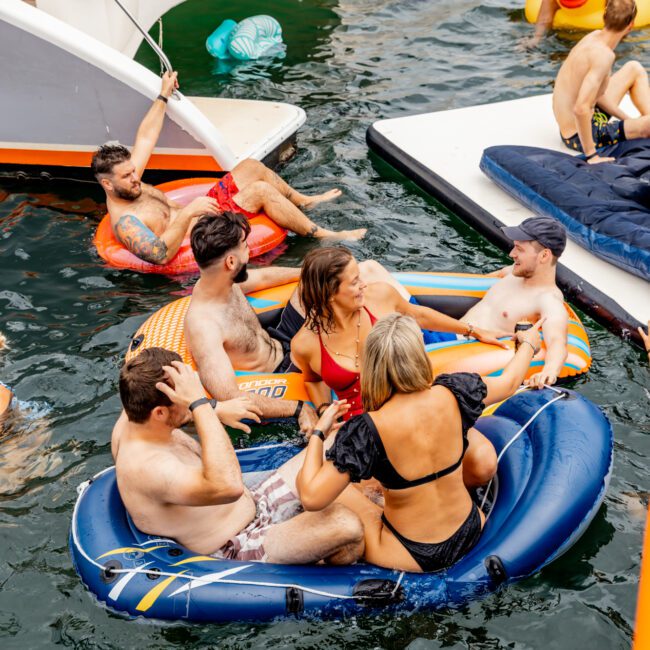 A group of people are relaxing on inflatable rafts in the water next to a boat. Some are chatting, others are enjoying the sun. The raft closest to the camera has three people, while another nearby has two. The environment at The Yacht Social Club Event looks festive and sociable.