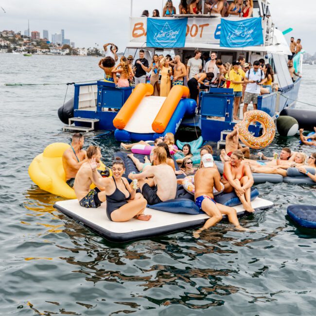 A large group of people in swimsuits are enjoying a lively party on The Yacht Social Club Sydney Boat Hire and surrounding inflatable platforms in the water. They are socializing, sunbathing, and swimming. The yacht has banners, and the backdrop features city buildings and a cloudy sky.