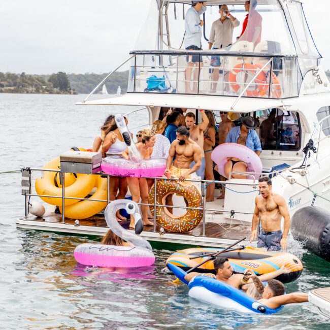 A lively scene unfolds as a group of people enjoy a summer day aboard The Yacht Social Club Sydney Boat Hire. Some are on the upper deck, while others lounge on the lower deck with inflatable floats shaped like donuts, swans, and a unicorn. A few float in the water on inflatable tubes, reveling in the moment.