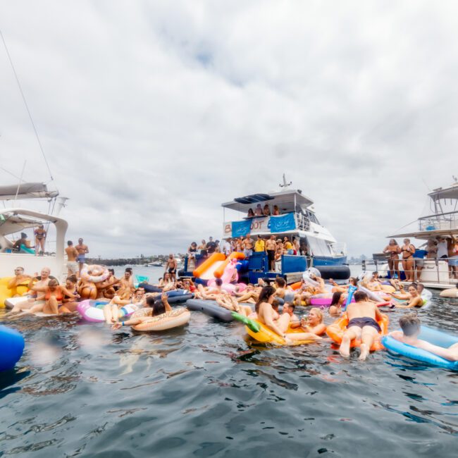 A large group of people in swimsuits enjoy a lively gathering on an array of inflatable rafts and floaties in the water, surrounded by several anchored boats. The atmosphere appears festive and vibrant, reminiscent of The Yacht Social Club Event Boat Charters.