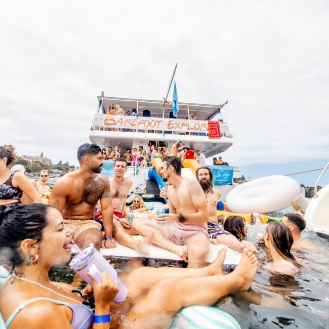 A group of people in swimwear enjoy a lively gathering on "Barefoot Explorer" and in the water alongside it. Some are on inflatables, and others are on the boat. They smile, laugh, and chat under a cloudy sky during one of the exclusive Sydney Harbour Boat Hire The Yacht Social Club events.