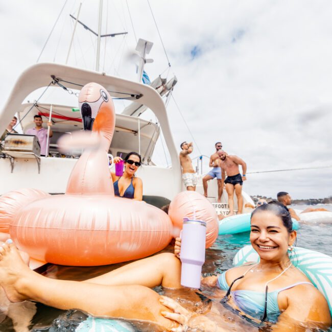 A group of people enjoying a sunny day on a boat and in the water. One woman in the foreground is smiling while sitting in a striped inflatable tube and holding a drink. Another person floats nearby in a pink flamingo inflatable. The Yacht Social Club Event Boat Charters hosts the perfect luxury yacht experience with The Yacht Social Club.