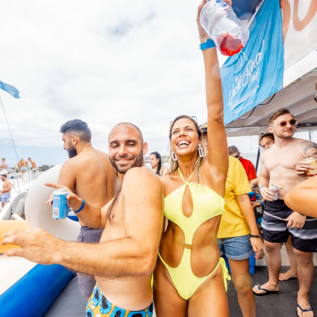 A group of people enjoys a party on a boat. In the foreground, a woman in a yellow swimsuit holds a drink and raises her arm, smiling, while standing with a man in blue patterned swim trunks. Others in the background also hold drinks and socialize at The Yacht Social Club Sydney Boat Hire event.