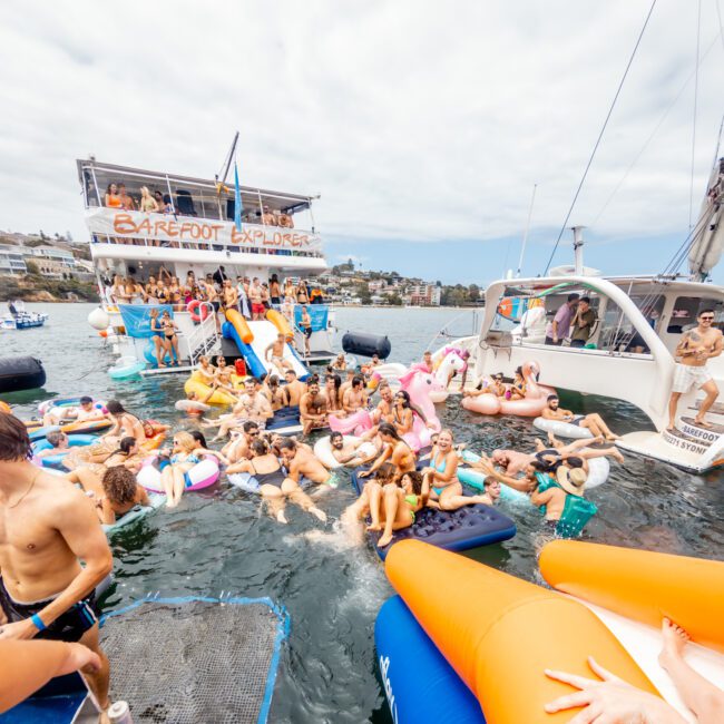 A large group of people are having a lively party in the water, surrounded by inflatables and boats. Many are wearing swimsuits, and some are on large floaties with colorful shapes. In the background, there's a boat named "Barefoot Explorer" docked nearby, offering a glimpse into The Yacht Social Club Event Boat Charters.