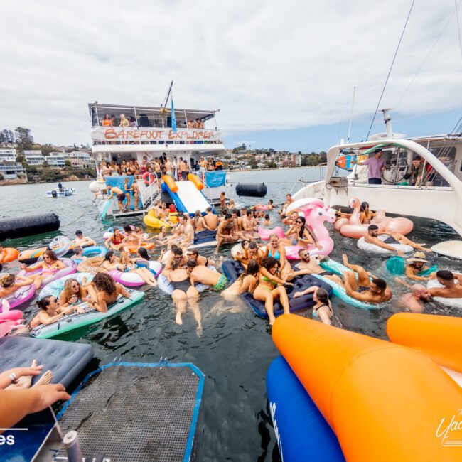 A lively scene of people enjoying a floating party in a marina. Many individuals are on colorful inflatable floats in the water. In the background, there is a large boat with “Barefoot Explorer” written on its side, part of The Yacht Social Club Event Boat Charters, and houses on a hill are visible.