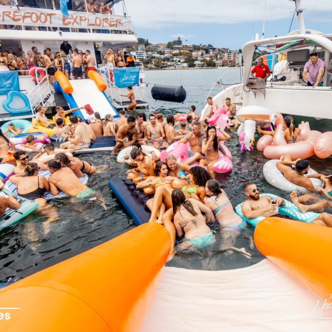 A lively scene of numerous people enjoying a floating party in a waterway. Attendees are relaxing on various inflatable devices, with a docked boat from The Yacht Social Club Sydney Boat Hire in the background. The atmosphere is vibrant with lots of social interaction.