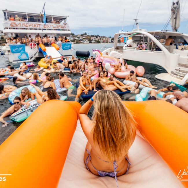 A lively boat party is taking place on the water. People are lounging on various inflatable float toys, including an orange slide and a pink unicorn. A large boat with "Barefoot Explorer" written on it is in the background. The scene, reminiscent of The Yacht Social Club Sydney Boat Hire gatherings, is energetic and festive.