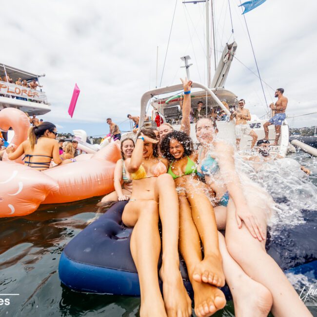 A group of young adults in swimsuits enjoy a party on the water. Some are sitting on a large float while others splash water nearby. The backdrop includes boats, inflatables, and people lounging. The atmosphere is lively and festive aboard a luxury yacht rented through Sydney Harbour Boat Hire.