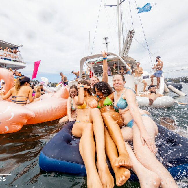 A group of friends in swimwear are lounging on a floating mat in the water, surrounded by inflatables like a pink flamingo and a unicorn. Nearby, boats from Sydney Harbour Boat Hire The Yacht Social Club are anchored. Everyone appears to be enjoying a sunny day on the water.