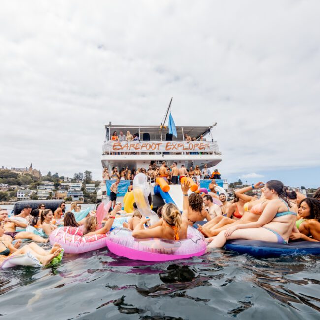 A festive scene on the water features a group of people on colorful inflatable rafts and floaties, enjoying a sunny day. In the background, "The Yacht Social Club Event Boat Charters" is filled with more people. Clear skies and waterfront buildings complete the lively atmosphere.