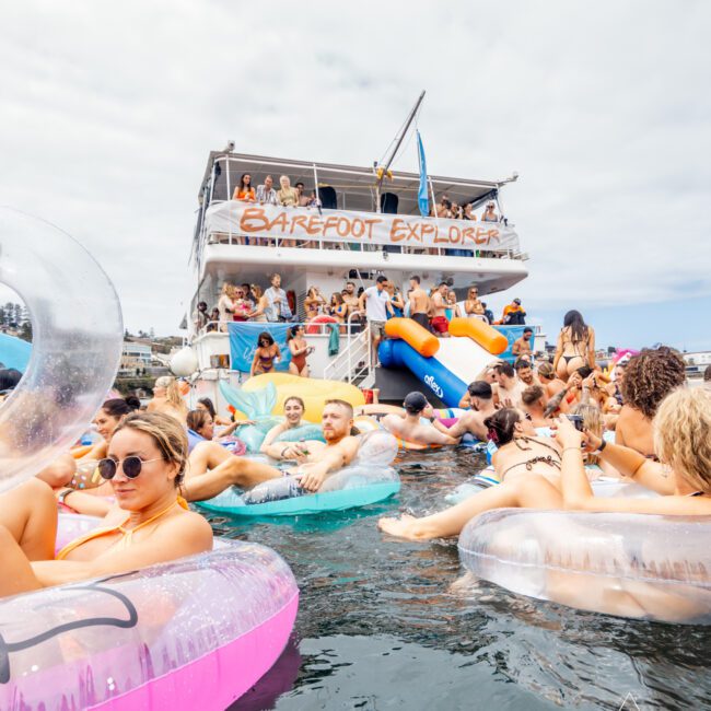 A lively pool party scene features many people relaxing on inflatable floats in the water. In the background, a double-decker boat with a sign reading "BAREFOOT EXPLORER" is docked. The atmosphere is festive and buzzing, reminiscent of Sydney Harbour Boat Hire The Yacht Social Club events.