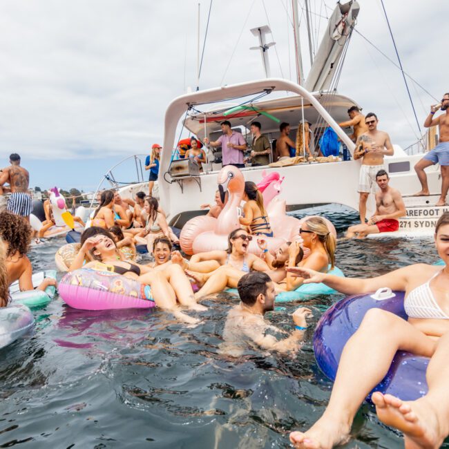 A lively group of people are enjoying a party on the water, floating on various inflatables near boats. They're smiling, laughing, and socializing under a cloudy sky. The scene is festive and relaxed with many in swimsuits and summer attire at The Yacht Social Club event.