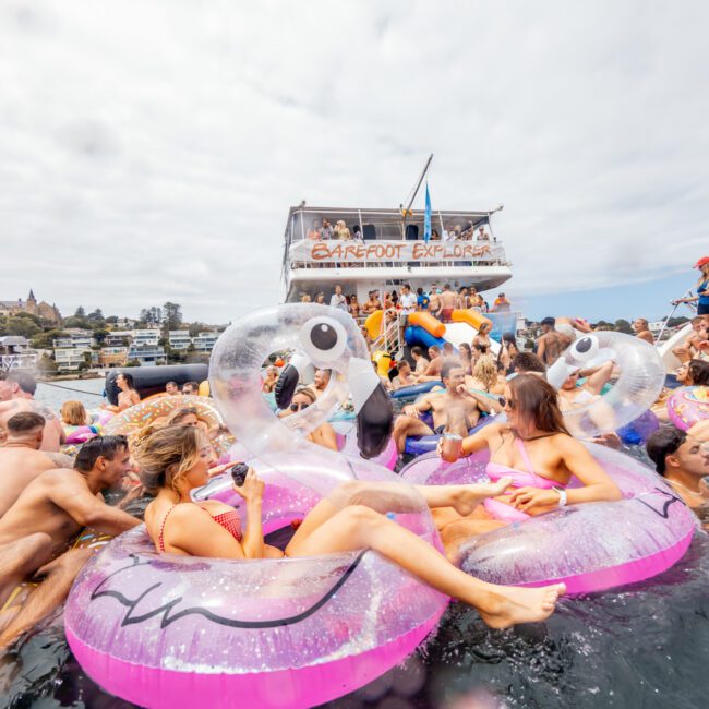 People enjoying a lively party in the water, surrounded by numerous inflatable floats, including pink flamingos. Boats are in the background, and a banner reading "BARFOOT EXPLORER" is visible. The sky is cloudy, adding a vibrant atmosphere to this fun-filled Yacht Social Club event.