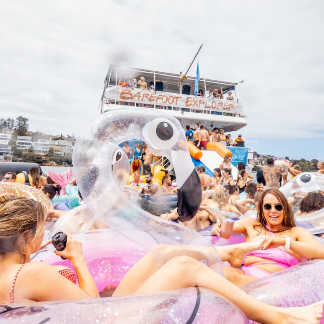 A large group of people enjoying a boat party, with many sitting on colorful inflatables in the water. A woman in a pink swimsuit is lying on a pink flamingo float, smiling and holding a drink. In the background, "Barefoot Explorer" by Sydney Harbour Boat Hire is visible for Luxury Yacht Rentals Sydney.