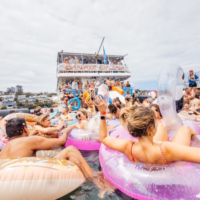 A lively scene of people enjoying themselves in a body of water, lounging on colorful inflatable floats. A two-story boat named "Barefoot Explorer" is anchored in the background, with more people on board. In the corner, a logo reads "The Yacht Social Club Sydney Boat Hire.