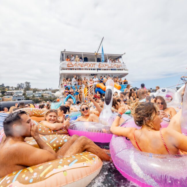 A lively party is taking place on a boat surrounded by people in the water on various inflatable floaties, including swans and doughnuts. The boat, part of The Yacht Social Club Sydney Boat Hire, has a sign that reads "Barefoot Explorer" and is filled with people dancing and cheering under bright and sunny weather.