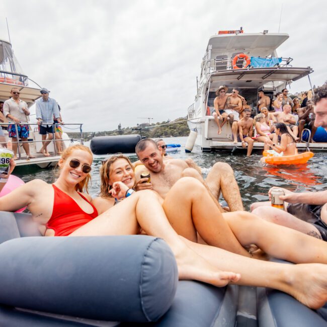 A group of people in swimsuits relax on inflatable lounges in the water, holding drinks and smiling at the camera. Behind them, others are enjoying themselves on two large boats anchored nearby. The atmosphere is lively and festive—typical of Boat Parties Sydney The Yacht Social Club.