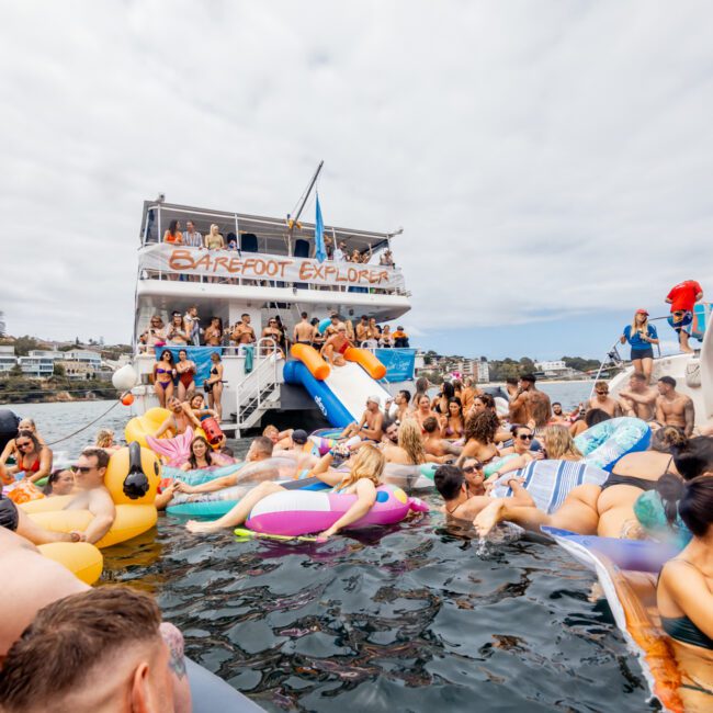 A lively party scene on the water with numerous people swimming and lounging on inflatable floats. Two boats are moored close by, one bearing the banner "BAREFOOT EXPLORER." The sky is overcast, and some attendees are on the boats' decks. Experience this with The Yacht Social Club Sydney Boat Hire.