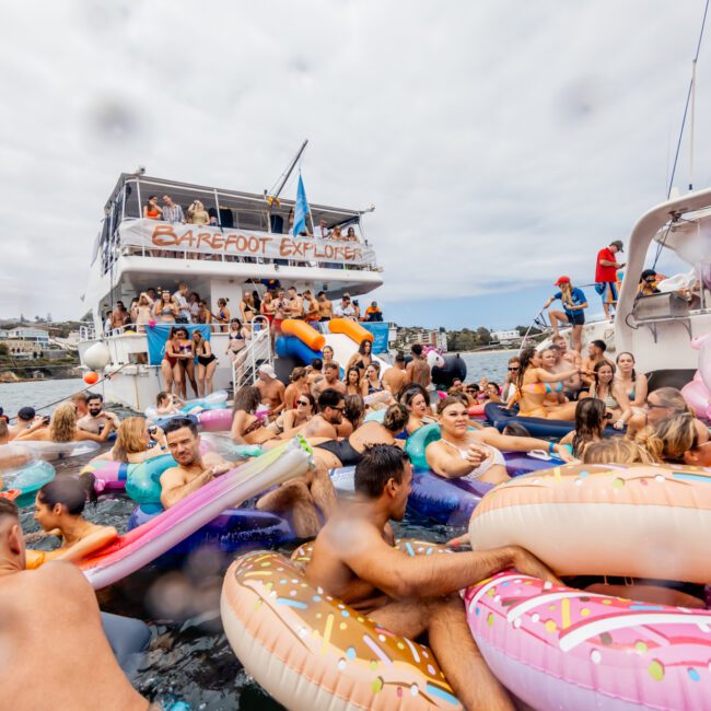 A lively crowd enjoying a boat party from The Yacht Social Club Sydney Boat Hire, with many floating on inflatable toys like doughnuts, a unicorn, and a flamingo in front of the double-decker boat. The atmosphere is festive, with coastal homes in the background.