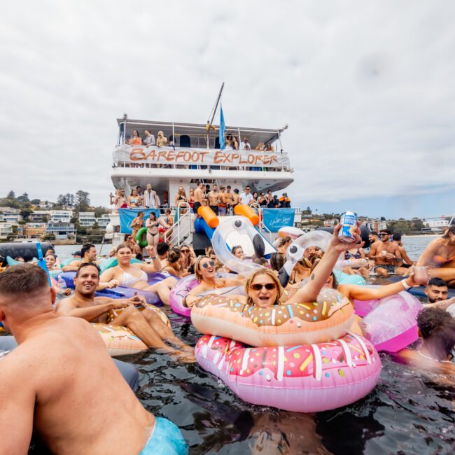 A large group of people enjoying a lively boat party with The Yacht Social Club. Many are on colorful inflatable rafts in the water, and a double-decker boat with "Barefoot Explorer" written on the banner is in the background. The atmosphere is festive and cheerful under a cloudy sky.