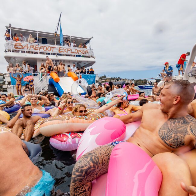 A lively scene of people gathered for a floating party with inflatable rafts and pool toys. A multi-level boat with a "Barefoot Explorer" banner is in the background, accompanied by houses lining the shore. The festive atmosphere comes alive with Luxury Yacht Rentals Sydney enhancing the vibrant event.