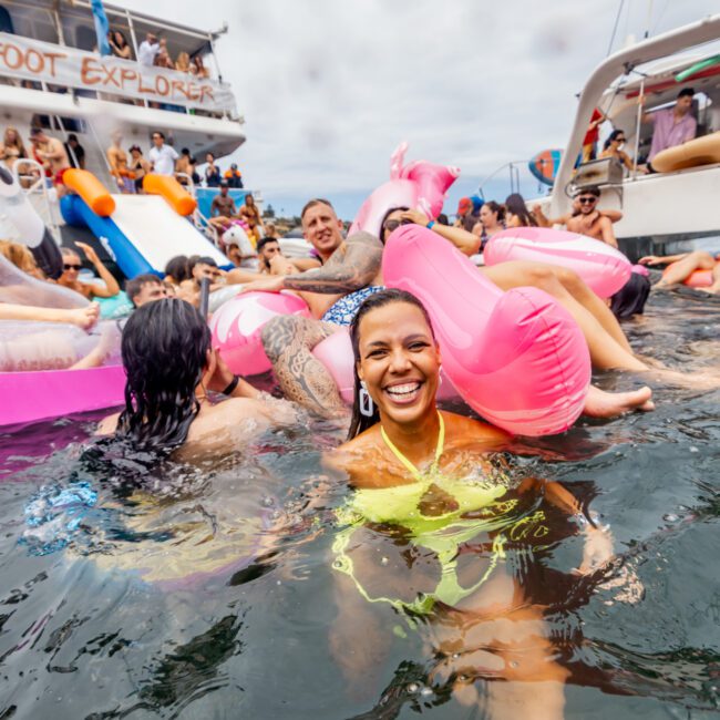 A joyful woman in a yellow swimsuit smiles among a colorful crowd on inflatable floats in the water. A party boat from The Yacht Social Club Sydney Boat Hire and another vessel are visible in the background with more people enjoying the festive atmosphere. The sky is overcast.