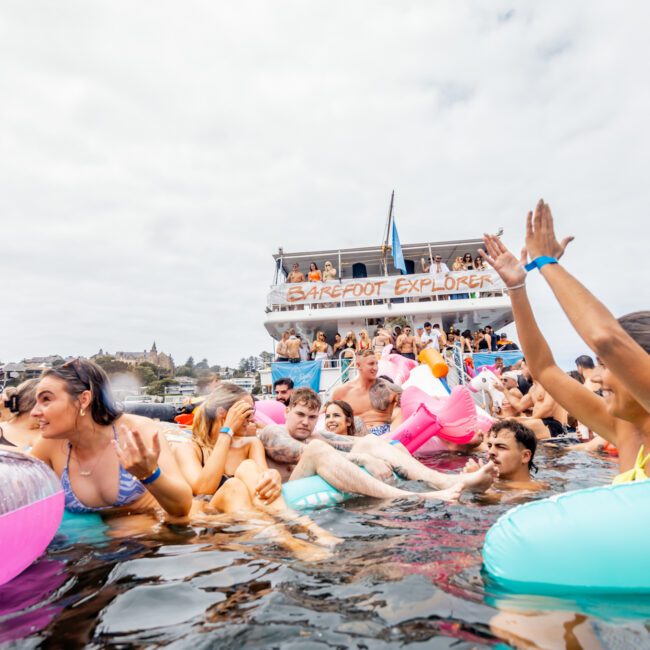 A lively scene of people enjoying a pool-like setting on the water with colorful floaties. The Yacht Social Club Event Boat Charters are in the background, filled with partying individuals. The atmosphere is festive with friends laughing, swimming, and raising their hands.