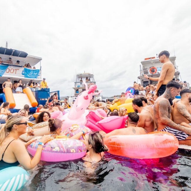 A lively gathering on the water with numerous people enjoying themselves on large inflatable floats. There are pink flamingo and unicorn floats among others. Two boats, provided by The Yacht Social Club Sydney Boat Hire, are anchored nearby, filled with more partygoers. The atmosphere is festive and energetic.