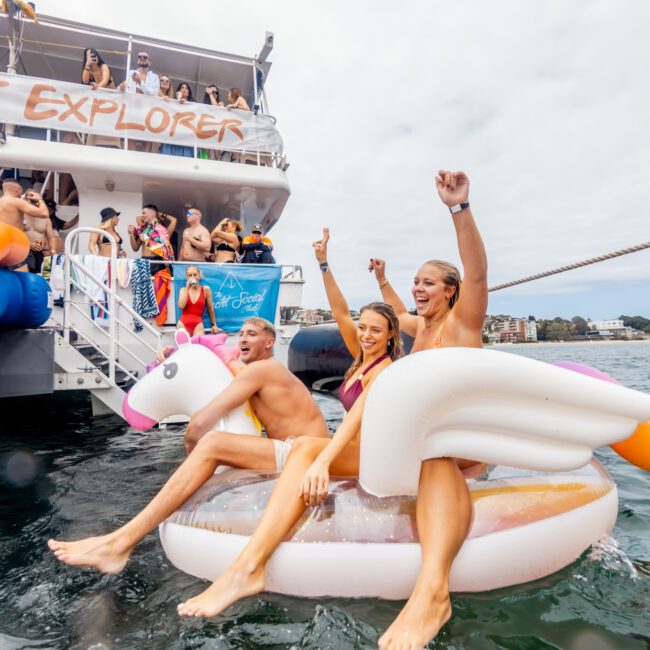 A group of young adults are enjoying a party on a boat named "Party Explorer." Three people are sitting on inflatable pool floats in the water—one on a unicorn float and another on a flamingo float. Others are on the boat, cheering and having fun. It’s another sensational event by The Yacht Social Club Sydney Boat Hire.