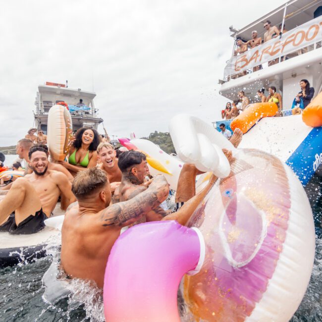 A group of people enjoys a lively party on the water, sitting on inflatable floaties and laughing. Some are sliding down a large water slide from a boat named "BAREFOOT" in the background. Hosted by The Yacht Social Club Sydney Boat Hire, the scene is vibrant with colorful floaties and everyone having a fun time.