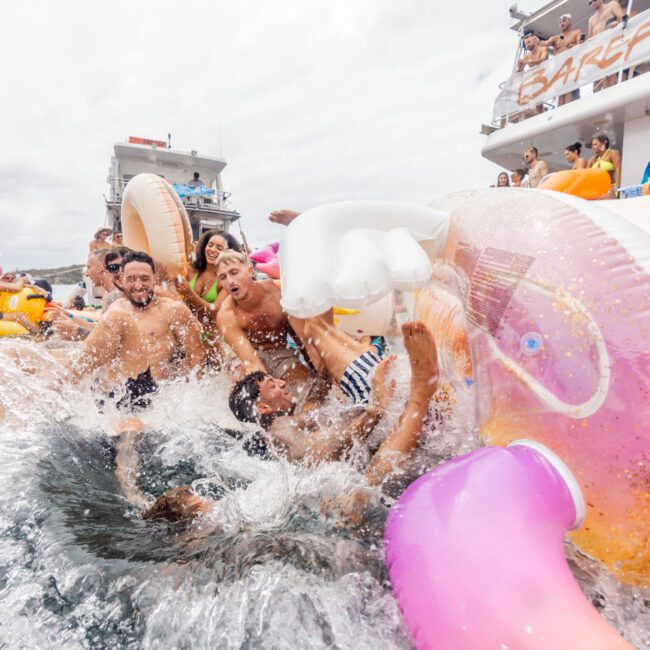 A group of people are having fun in the water near several boats. They are laughing and splashing, with some on large inflatable floaties shaped like unicorns and other bright designs. The sky is overcast, and the mood is lively and joyful at The Yacht Social Club Sydney Boat Hire.