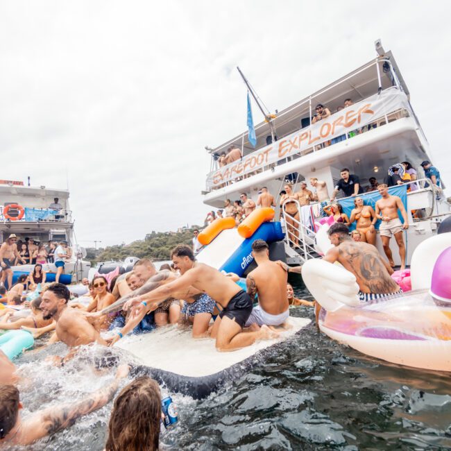 A lively scene unfolds at The Yacht Social Club Sydney Boat Hire, where people enjoy inflatables and swim around two anchored boats. The boats are filled with attendees, some jumping into the water. Inflatables in various shapes, including unicorns, add to the fun. The sky is cloudy.