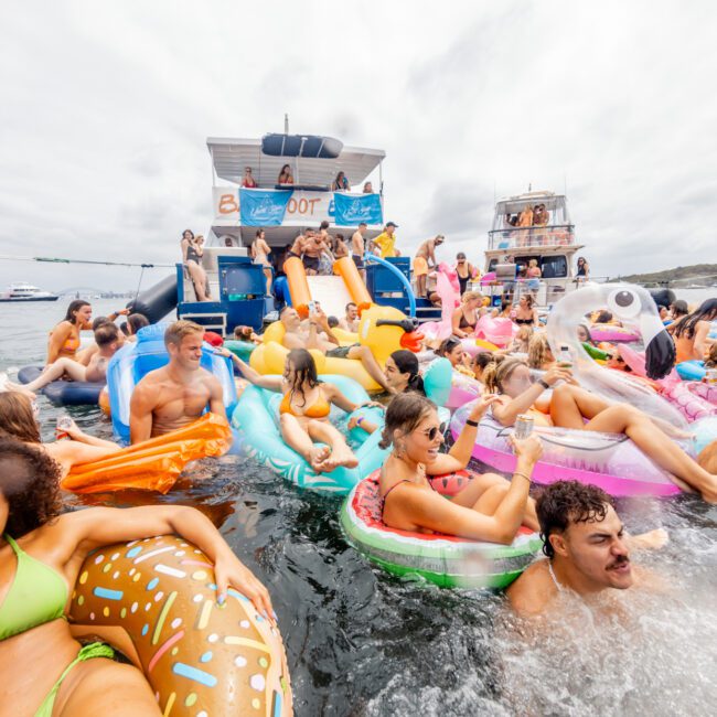 A lively group of people enjoys a boat party, relaxing on colorful inflatable floats in the water. The scene features a double-decker boat from The Yacht Social Club Event Boat Charters with a slide, more partygoers on the upper deck, and extra boats in the background. Everyone seems to be having fun.