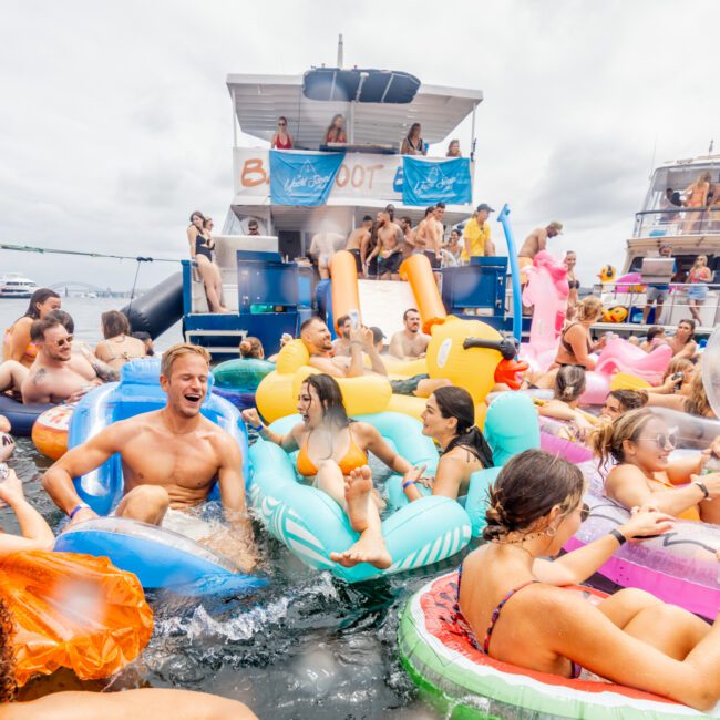 A large group of people enjoying a lively boat party with The Yacht Social Club, many on colorful inflatable floats in the water. The scene is festive, with individuals laughing, talking, and sliding down an inflatable slide from the boat's deck. The sky is cloudy.