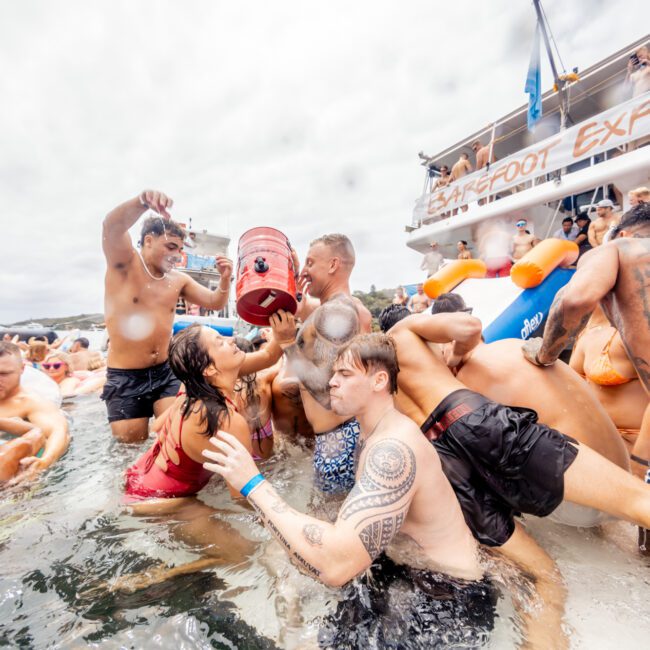 A group of people in swimwear are gathered closely together in water, participating in a lively event. Some are taking drinks from a large jug. There is a boat labeled 'Sloopfoot Express' in the background, part of The Yacht Social Club Sydney Boat Hire, and everyone appears to be having an energetic and festive time.