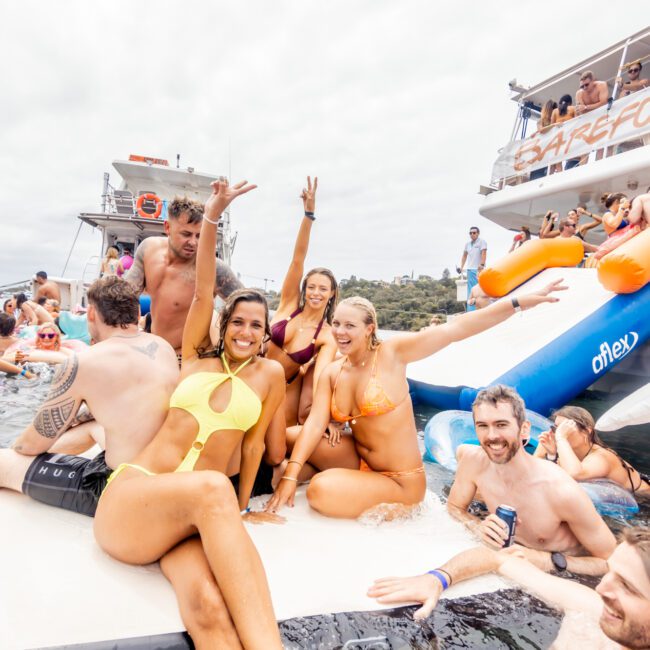 A group of people in swimsuits enjoy a party on the water, with some sitting on a float and others in the water. They are smiling, holding drinks, and posing for the camera. A large inflatable slide and anchored yachts are visible in the background—an iconic scene from Sydney Harbour Boat Hire The Yacht Social Club.