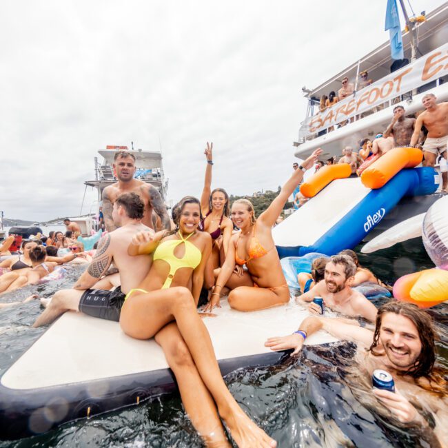 A lively group enjoys a boat party on the water. Some are on a float, while others swim around with inflatable pool toys, including a duck. A large boat with a water slide is in the background. People are smiling and waving. Hosted by The Yacht Social Club, it's a perfect example of Boat Parties Sydney's finest moments.